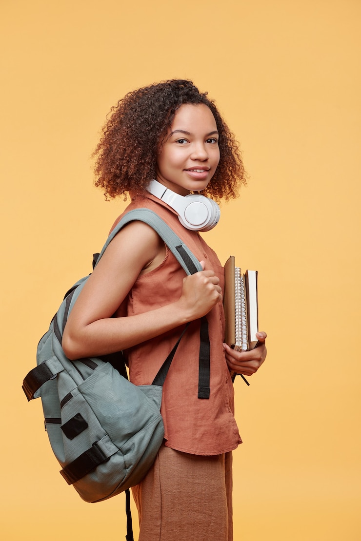 Foto portret van positief mooi studentenmeisje met krullend haar die schooltas op rug met stapel werkmappen dragen tegen gele achtergrond