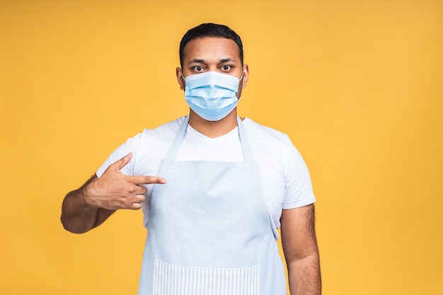 Portret van positieve aantrekkelijke afro-amerikaanse zwarte indiase kok man in schort en beschermend masker kijken camera geïsoleerd op gele achtergrond. coronavirus-concept.