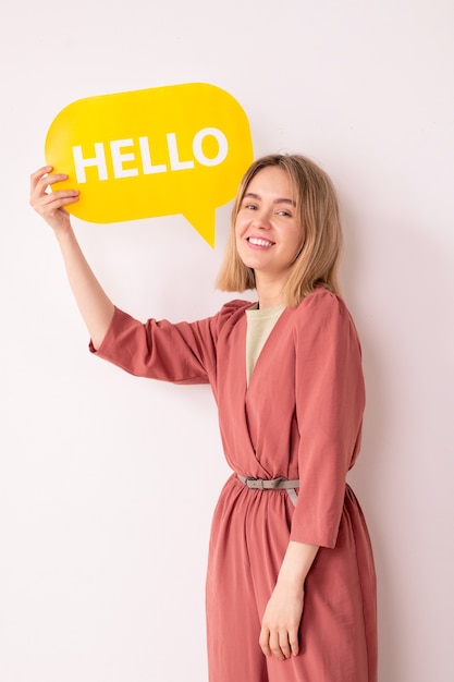 Portret van positieve aantrekkelijke jonge vrouw in roze jumpsuit die communicatie-tag toont terwijl ze hallo zegt