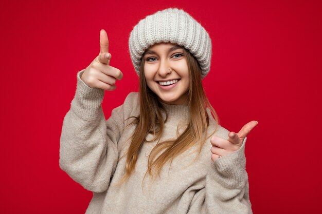 Portret van positieve gelukkig lachende jonge mooie donkere blonde dame met oprechte emoties dragen beige trui en gebreide muts geïsoleerd op rode achtergrond met kopie ruimte en wijzend op de camera