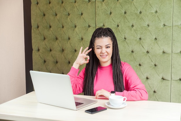 Portret van positieve grappige jonge vrouw met zwarte dreadlocks kapsel in roze blouse zitten in café en tonen ok teken met hand knipogen camera en brede glimlach Indoor levensstijl