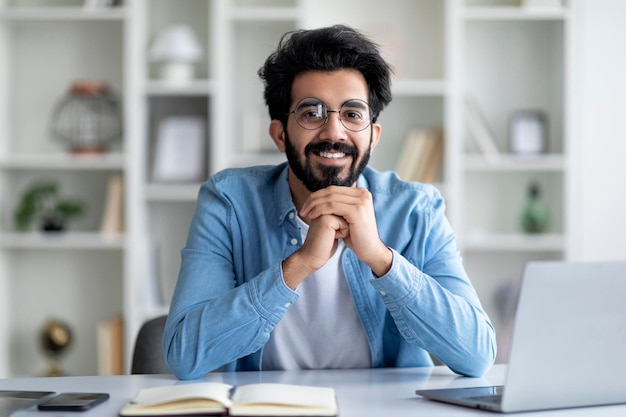 Portret van positieve Indiase freelancer man zit aan bureau in kantoor aan huis