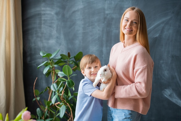 Portret van positieve jonge moeder en haar zoon poseren met konijn tegen bord