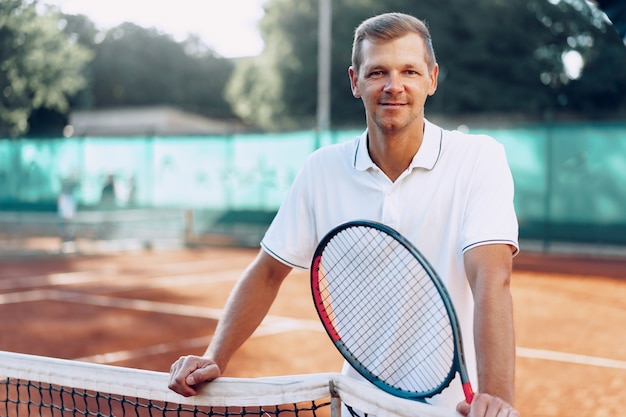 Portret van positieve mannelijke tennisser met racket staande op gravel in de buurt van net