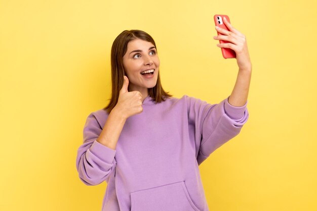 Portret van positieve mooie vrouw die hand naar smartphone zwaait en selfie neemt met duim omhoog terwijl ze videogesprek voert met paarse hoodie Indoor studio shot geïsoleerd op gele achtergrond