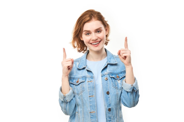 Portret van positieve roodharige jonge vrouw in casual jeans glimlachend wijzend met de vinger naar lege kopie ruimte voor tekst of product geïsoleerd op een witte achtergrond reclamebanner