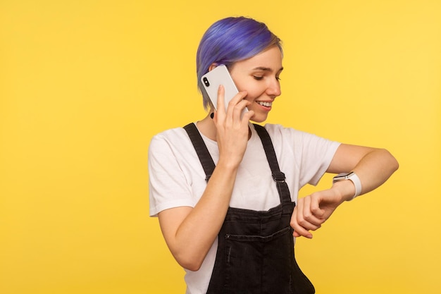 Portret van positieve schattige hipster meisje met violet kort haar in overall praten op mobiele telefoon en kijken naar horloge controleren tijd haasten op vergadering geïsoleerd op gele achtergrond studio opname