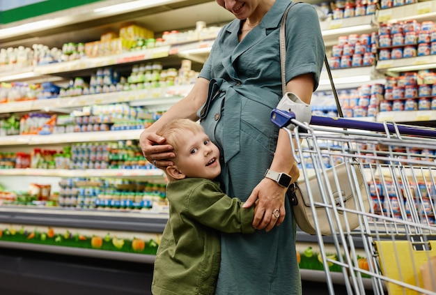 Portret van positieve schattige zoon met blond haar die zwangere moeder omhelst in supermarkt