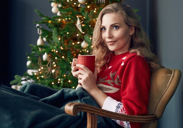 Portret van prachtige blonde vrouw in rode trui koelen onder een plaid met een kopje koffie in het kerst versierde interieur.