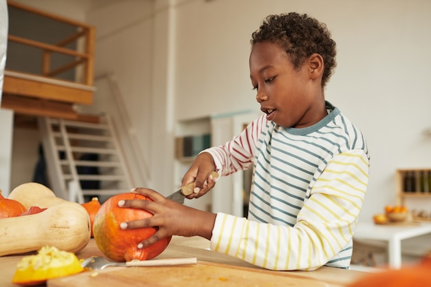 Portret van preteen afro-amerikaanse jongen, gekleed in casual outfit staande aan tafel in keuken pompoen snijden voor halloween