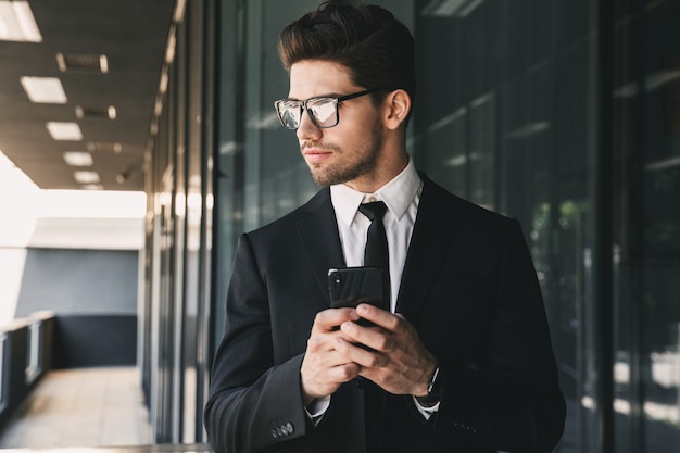 Portret van professionele jonge zakenman gekleed in formeel pak staande buiten glazen gebouw, en smartphone te houden