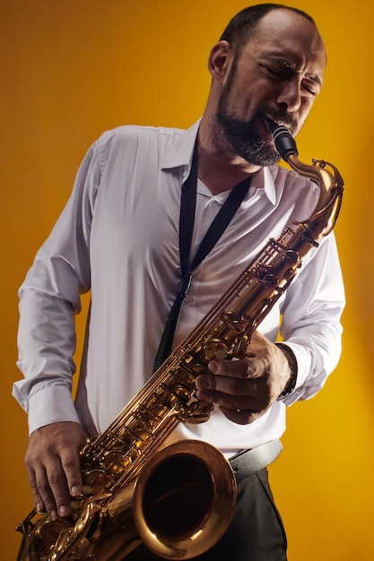 Portret van professionele muzikant saxofonist man in wit overhemd speelt jazzmuziek op saxofoon, gele achtergrond in een fotostudio, zijaanzicht