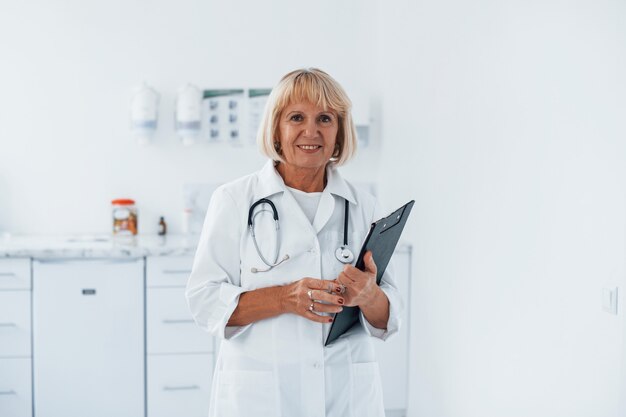 Foto portret van professionele senior arts in kliniek met kladblok in handen.