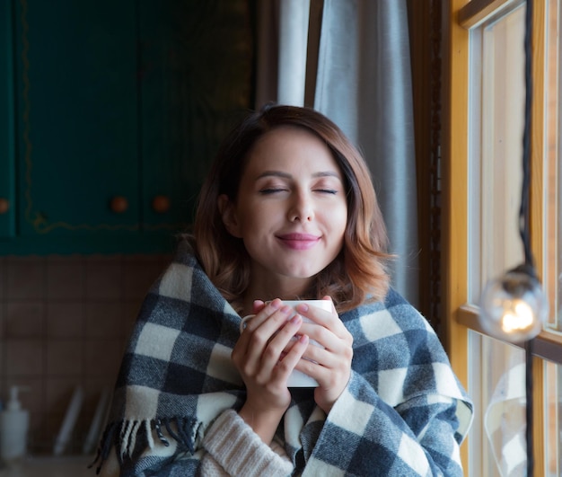 Portret van roodharige vrouw met plaid en kopje koffie bij het raam, gezellig ochtendconcept