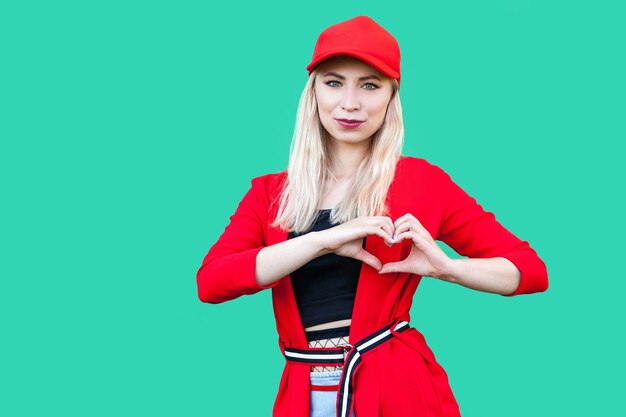 Portret van rustige mooie blonde jonge hipster vrouw in rode blouse en pet, permanent met hart hand teken gebaar en kijken naar camera en glimlach. indoor studio opname, geïsoleerd op groene achtergrond.