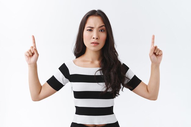 Portret van sceptische onzekere brunette Oost-Aziatische vrouw in gestreepte tshirt fronsen en het verhogen van een wenkbrauw loensen met ongeloof wijzende vingers omhoog uitdrukkelijke twijfels witte achtergrond