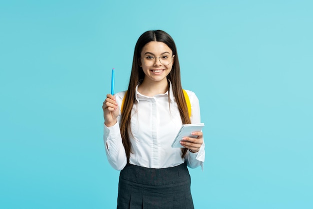 Portret van schattig glimlachend tienermeisje met bril en schooluniform schrijven in notitieboekje geïsoleerd op blauwe achtergrond Schoolconcept