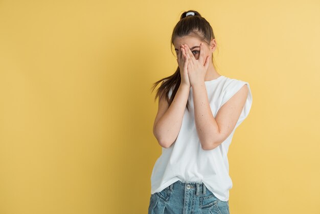 Portret van schattig jong Europees meisje gezicht met hand kijken camera door vingers voor gele studio achtergrond verbergen.