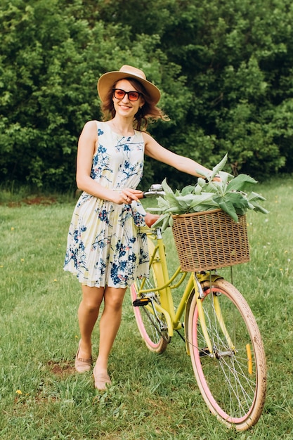 Portret van schattig meisje met krullend blond haar in hoed een fiets rijden. Actieve mensen. Buitenshuis. Mooi meisje op fiets in platteland ruikende bloemen, zomer levensstijl. close-up portret