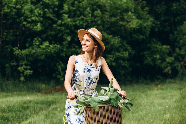 Portret van schattig meisje met krullend blond haar in hoed een fiets rijden. Actieve mensen. Buitenshuis. Mooi meisje op fiets in platteland ruikende bloemen, zomer levensstijl. close-up portret