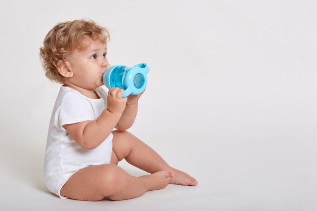 Portret van schattig peuter drinkwater uit de fles zittend tegen een witte muur, pak dragen