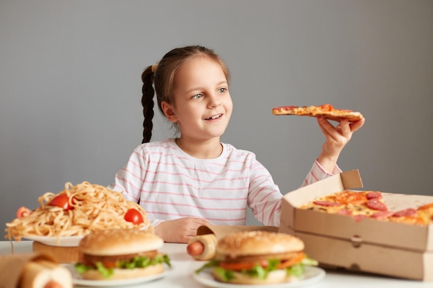 Portret van schattig schattig klein meisje met staartjes zittend aan tafel geïsoleerd over grijze achtergrond houden pizza slice eten fastfood poseren onder junk schotel