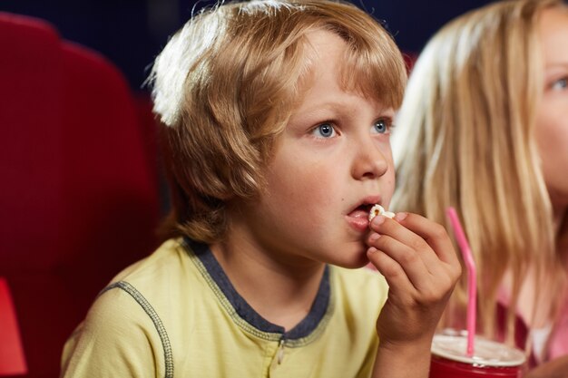 Portret van schattige blonde jongen kijken naar film in bioscoop theater en eten popcorn, kopie ruimte