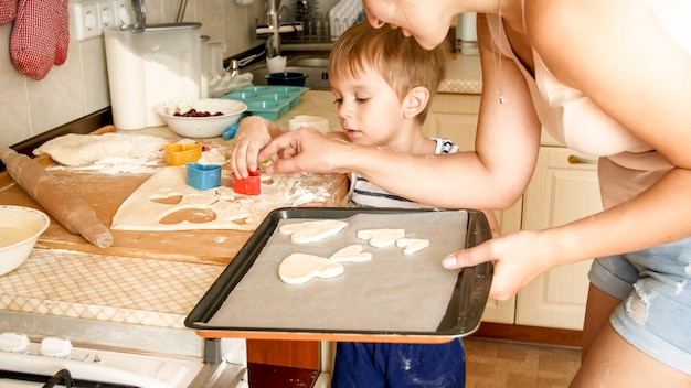 Portret van schattige kleine jongen met jonge moeder die koekjes bakt op bakpan in de keuken