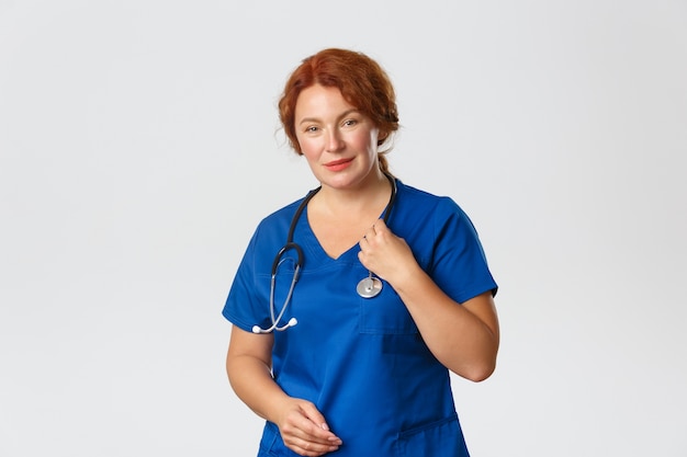 Portret van schattige roodharige vrouwelijke arts, verpleegster of arts in blauwe scrubs die patiënt met een stethoscoop controleren