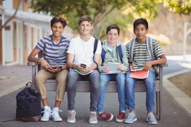 Portret van schoolkinderen met behulp van mobiele telefoon en digitale tablet op bankje op schoolcampus