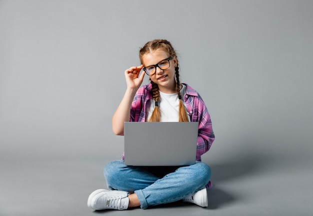 Portret van schoolmeisje meisje met laptop zittend in kleermakerszit geïsoleerd op een grijze achtergrond