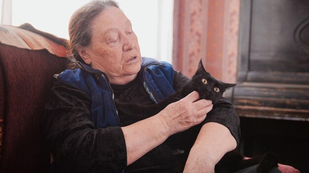 Portret van senior dame thuis - oude vrouw zit op de bank met zwarte kat, close-up, telefoto