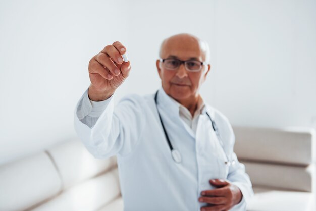 Portret van senior dokter wit uniform dat in de kliniek staat met pil in de hand.