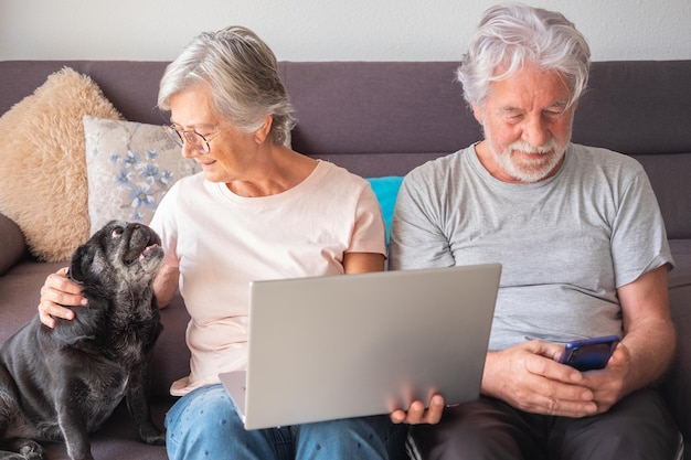 Portret van senior volwassen Kaukasisch koppel zittend op de bank met oude pug dog praten en plezier hebben samen met liefde met behulp van mobiele telefoon en laptop Concept van diertherapie voor bejaarde oude persoon