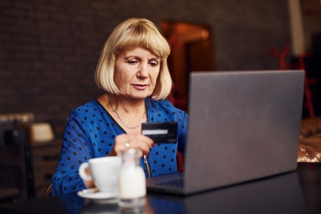 Portret van senior vrouw die binnen in het café zit met moderne laptop en creditcard in de hand.