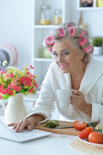 Portret van senior vrouw in badjas met krulspelden zittend aan tafel met laptop