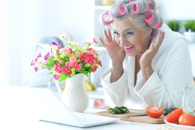 Portret van senior vrouw in badjas met krulspelden zittend aan tafel met laptop