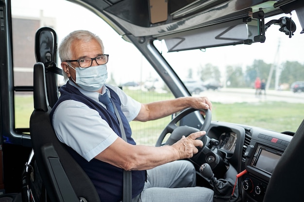 Portret van senior witharige buschauffeur in brillen en gezichtsmasker vastmaken veiligheidsgordel zittend aan ...