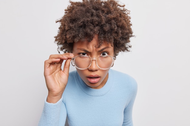 Portret van serieuze Afro-Amerikaanse vrouw met krullend haar kijkt aandachtig door transparante bril luistert iets heeft geërgerde uitdrukking gekleed in casual trui geïsoleerd over witte muur