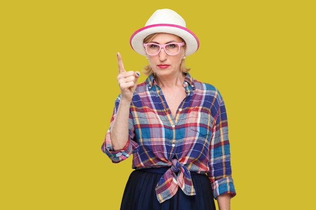 Portret van serieuze bazige moderne stijlvolle volwassen vrouw in casual stijl met hoed en bril die met een waarschuwingsbord staat en naar de camera kijkt. indoor studio opname geïsoleerd op gele achtergrond.