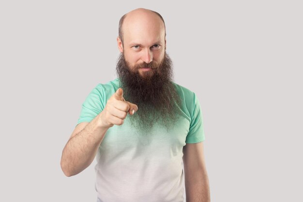 Portret van serieuze kale man van middelbare leeftijd met lange baard in lichtgroene tshirt permanent wijzend en kijkend naar camera binnen studio opname geïsoleerd op grijze achtergrond