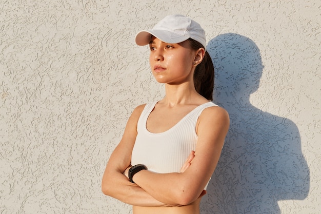 Portret van serieuze zelfverzekerde vrouw met witte top en vizierdop, geïsoleerd over grijze muur buiten met gevouwen handen, op afstand kijkend, sportieve vrouw na het sporten.