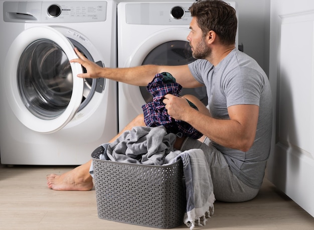 Portret van sexy man met vuile kleren in de buurt van wasmachine Knappe man zit voor wasmachine Vuile was Man kleren schoonmaken Huishoudelijk werk voor alleenstaande man Wasserij thuis