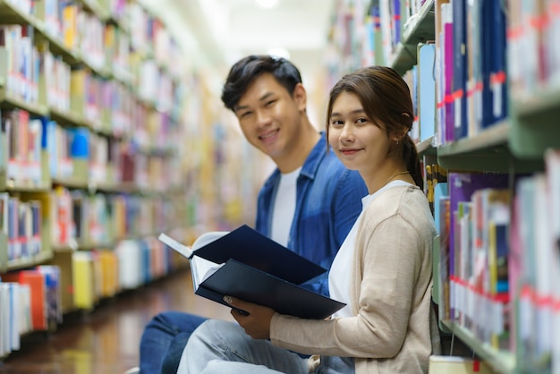 Portret van slimme Aziatische man en vrouw universitaire student leesboek samen en. camera kijken tussen boekenplanken in campusbibliotheek met copyspace.