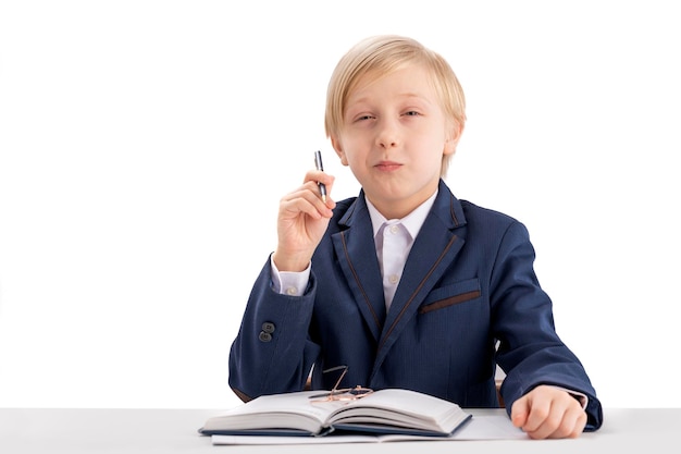 Portret van slimme blonde schooljongen in schooluniform student zit aan tafel met boek en pen in zijn handen onderwijsconcept isolatie