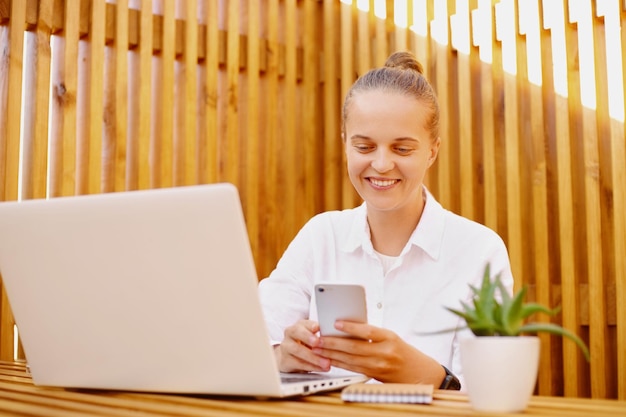 Portret van slimme jonge aantrekkelijke vrouw met mobiele telefoon die op laptop werkt op kantoor in de buitenlucht met een wit t-shirt dat haar e-mail controleert met mobiele telefoon in handen