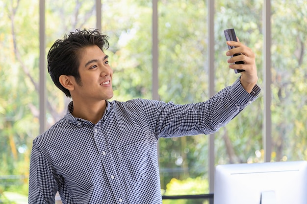 Portret van slimme jonge aziatische zakenman die slimme mobiele telefoon gebruikt om een selfie-foto te maken in de kantoorruimte.