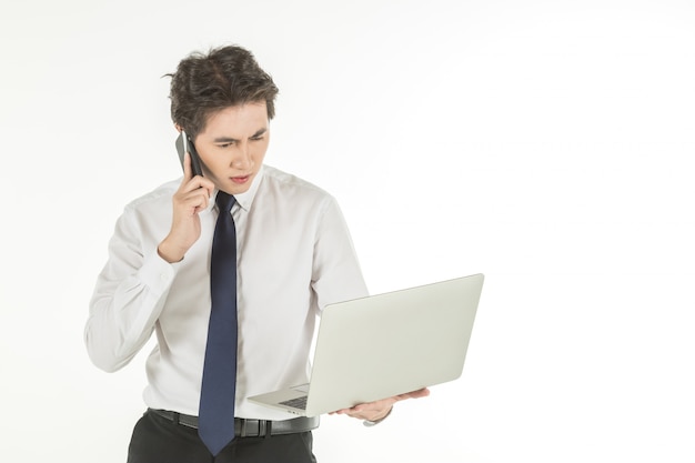 Portret van slimme jonge Aziatische zakenman die wit overhemd dragen die en aan computerlaptop houden kijken en met slimme mobiele telefoon telefoneren