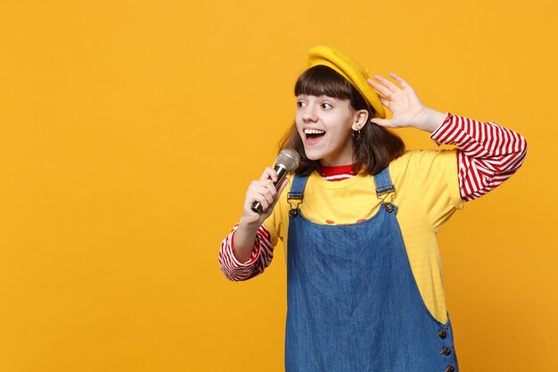 Portret van slimme tiener meisje in Franse baret, denim zomerjurk zingen lied in microfoon geïsoleerd op gele muur achtergrond in studio. Mensen oprechte emoties, lifestyle concept. Bespotten kopie ruimte.