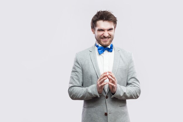 Portret van sluwe knappe bebaarde man in casual grijs pak en blauwe vlinderdas staande met grappig gezicht en palm hand camera kijken met idee. indoor studio opname, geïsoleerd op lichtgrijze achtergrond.